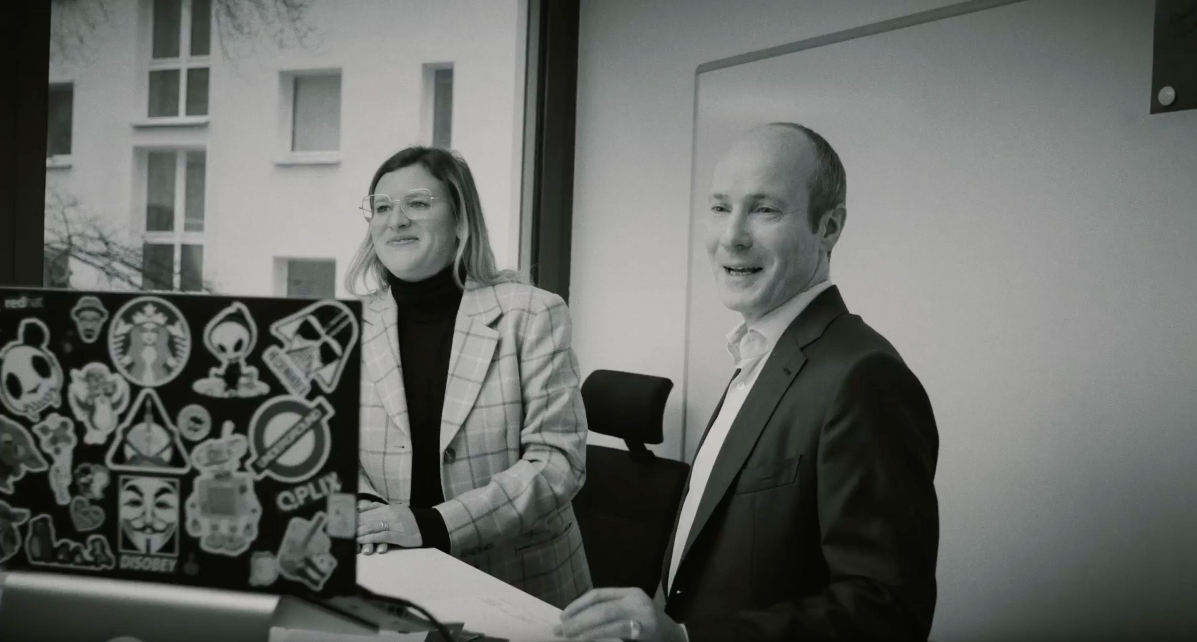 Schwarz-Weiß-Aufnahme zweiter Mitarbeitender stehend vor Tisch mit Laptop. Beide im Gespräch und lachend