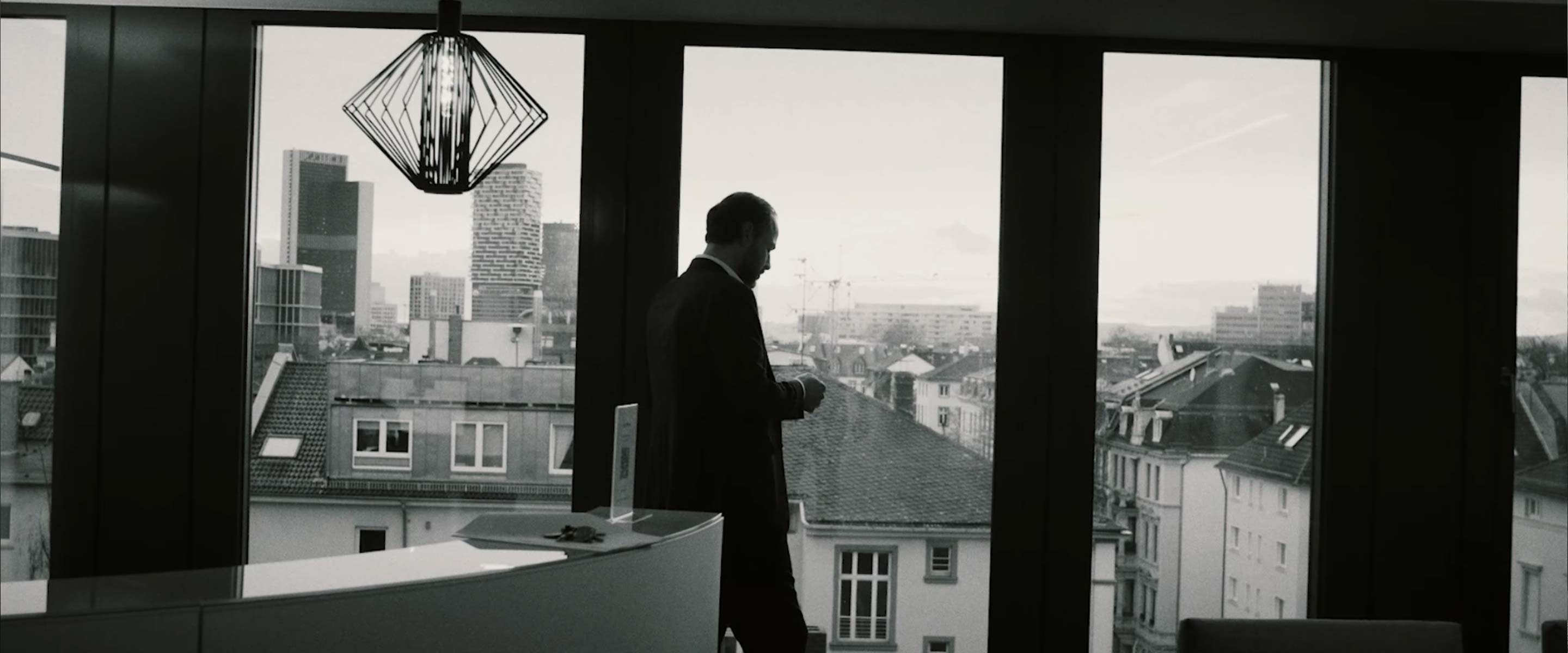 Schwarz-Weiß-Aufnahme. Im Hintergrund Ausblick auf Frankfurter Skyline aus großer Fensterfront. Im Vordergrund Mann im Anzug, der an Empfangstresen lehnt.