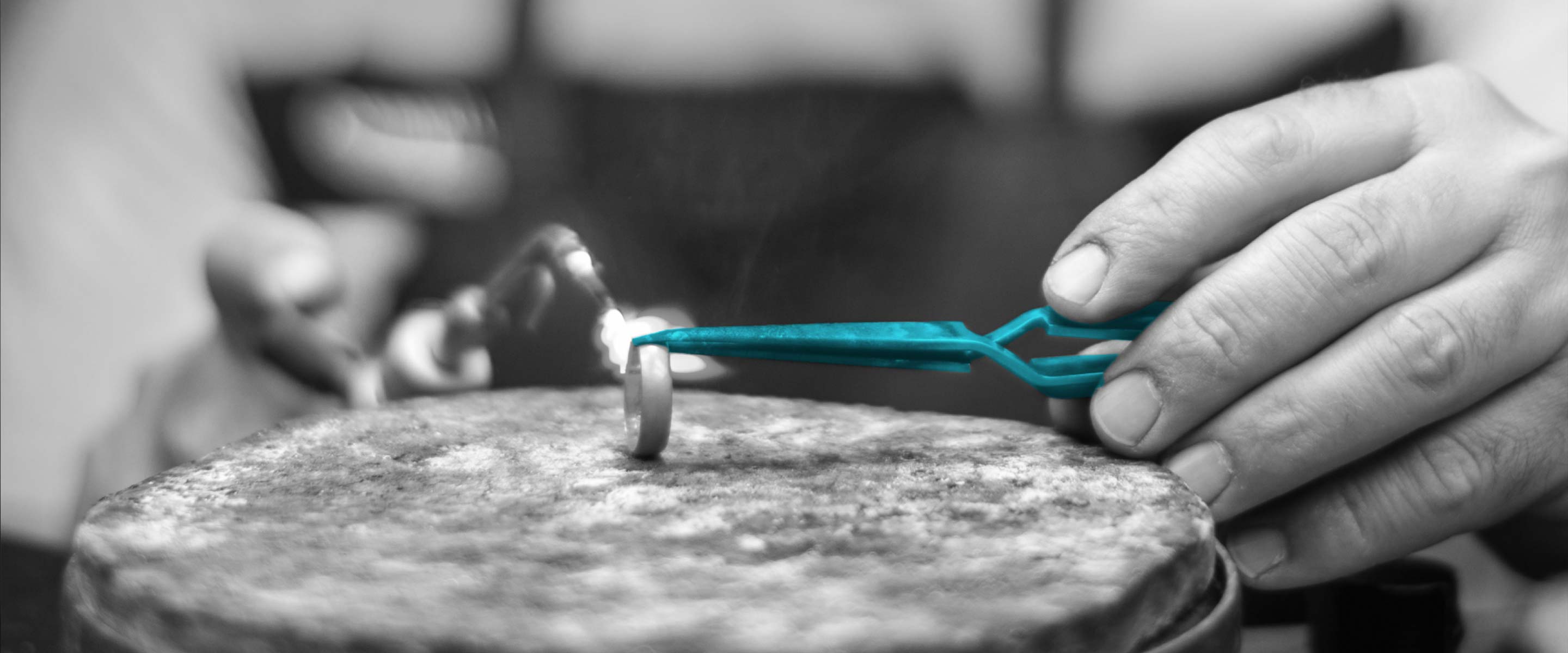 Schwarz-Weiß-Nahaufnahme der Bearbeitung eines Rings. Die Zange zu dessen Halt ist in petrol hervorgehoben