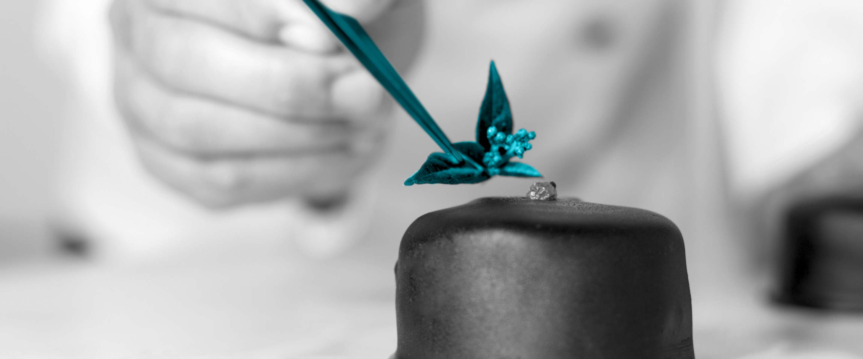 Nahaufnahme eines Juweliers bei der Arbeit mit Bildausschnitt der Hand. Bild in schwarz-weiß, nur Schmuckstück hervorgehoben in petrol